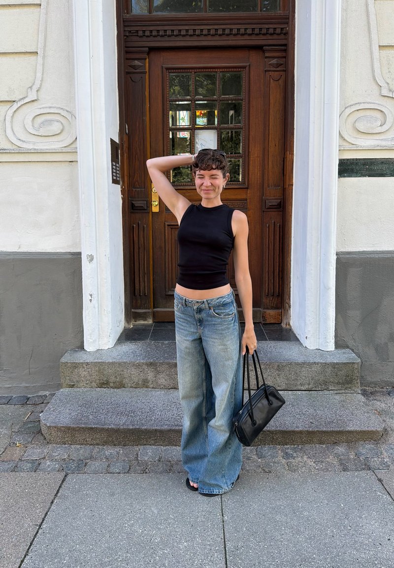 Junge Person mit kurzen lockigen Haaren, die ein schwarzes ärmelloses Top und weite Jeans trägt, steht auf Stufen vor einer Holztür.