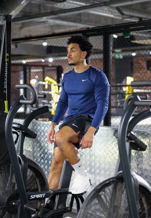 Hombre con camisa de manga larga azul y pantalones cortos negros sentado en una bicicleta estática en un gimnasio con decoración industrial.