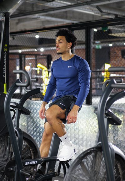 Hombre con camisa de manga larga azul y pantalones cortos negros sentado en una bicicleta estática en un gimnasio con decoración industrial.