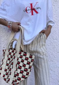 Hvid bomulds t-shirt med rød "Calvin Klein Jeans" logo, parret med beige stribede linnedbukser og en cremefarvet tote taske med et rødt og sort NY mønster.