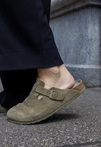 Brown suede clogs featuring a single buckle strap, an open back, and a textured sole. Paired with black trousers, highlighting the shoe's style and fit.