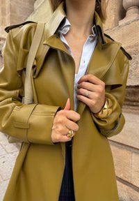 Ελαιόχρωμο δερμάτινο παλτό με μεγάλο γιακά, διπλό κούμπωμα και ορατό ράψιμο. Αξεσουάρ περιλαμβάνουν χρυσά δαχτυλίδια και ένα βραχιόλι.