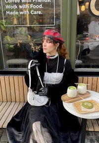 Ένα άτομο κάθεται σε ένα τραπέζι στον ύπαιθρο, φορώντας ένα μαύρο μπουφάν, ένα τοπ με δαντέλα και ένα καπέλο καρό. Στο τραπέζι υπάρχουν μια άσπρη τσάντα ώμου και ροφήματα matcha.