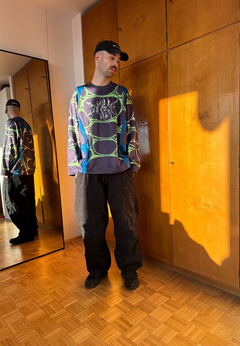 Hombre con gorra negra, camisa de manga larga estampada y pantalones negros holgados, de pie junto a unos armarios de madera reflejados en un espejo de cuerpo entero.