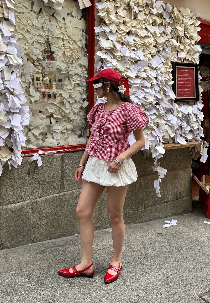 Blouse à manches bouffantes en damier rouge et blanc avec un détail à lacer, assortie à une jupe blanche à volants et des chaussures plates rouges brillantes avec une bride.