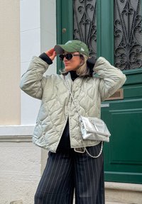 Veste matelassée gris clair, col roulé noir, pantalon à rayures noir et blanc, casquette verte, sac à bandoulière argenté avec chaîne. Porte verte en arrière-plan.