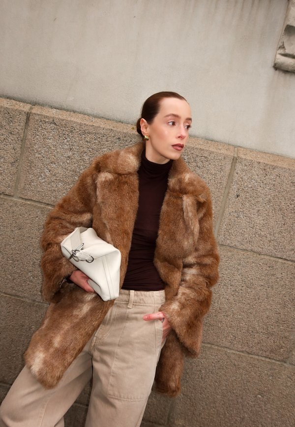 Vrouw in een bruine nepbontjas en beige broek houdt een witte clutch vast, terwijl ze tegen een stenen muur staat en naar de zijkant kijkt.