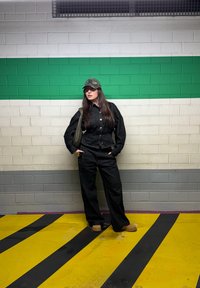 Femme en tenue en jean noir et chaussures marron, les mains dans les poches, se tenant contre un mur vert et blanc dans une zone de stationnement.