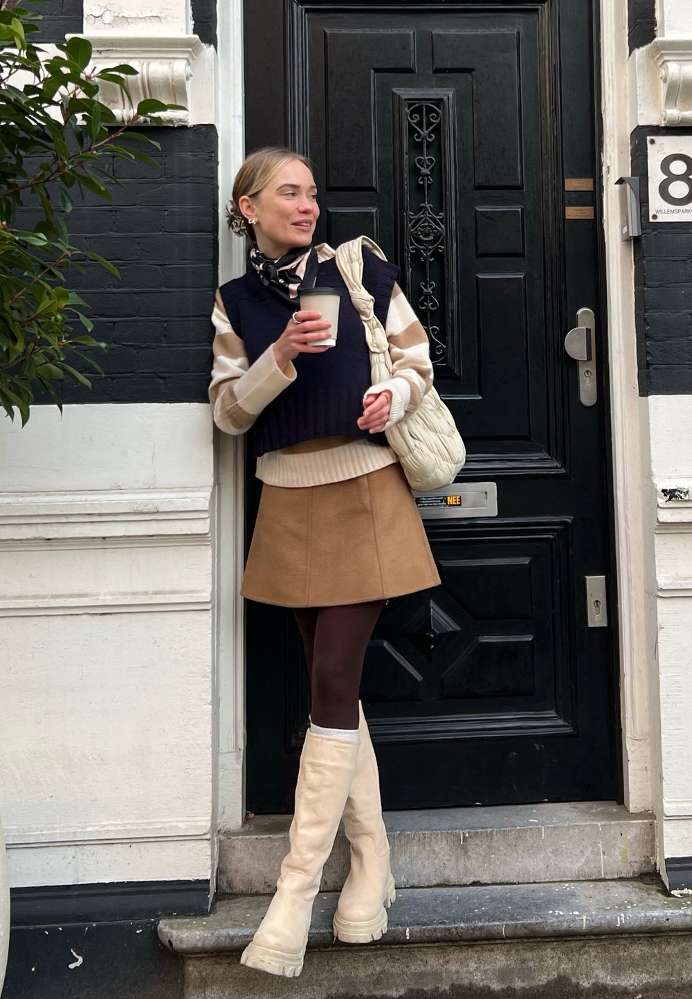 Botas bege até ao joelho, saia mini castanha, colete de sweater azul-marinho sobre uma blusa creme, segurando uma chávena de café, em pé junto a uma porta decorativa preta.
