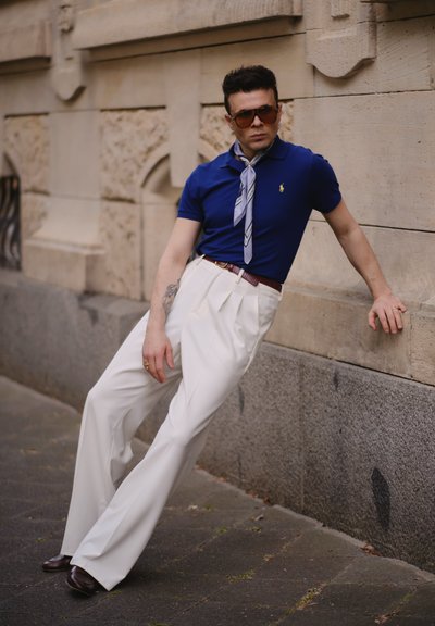 Modelo con una camisa polo azul con un pequeño logo, pantalones blancos con pliegues y una bufanda ligera de estampado, usando gafas de sol oscuras y zapatos marrones.