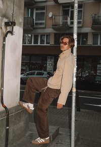 Gaiši bēša jaka, brūnas bikses un brūnas sporta kurpes ar baltām svītrām. Stāv uz pakāpiena, atspiežoties pret stabu pilsētas vidē.