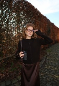 Femme en cardigan noir et jupe marron tenant une tasse de café et ajustant ses lunettes, debout sur un chemin pavé à côté d'une haie d'automne haute.