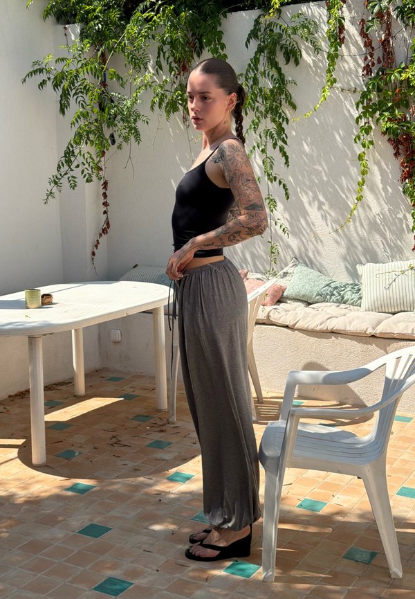 Ung kvinde med tatoveringer iført sort tanktop og grå bukser står ved siden af en hvid plastikstol og bord på en solrig terrasse.