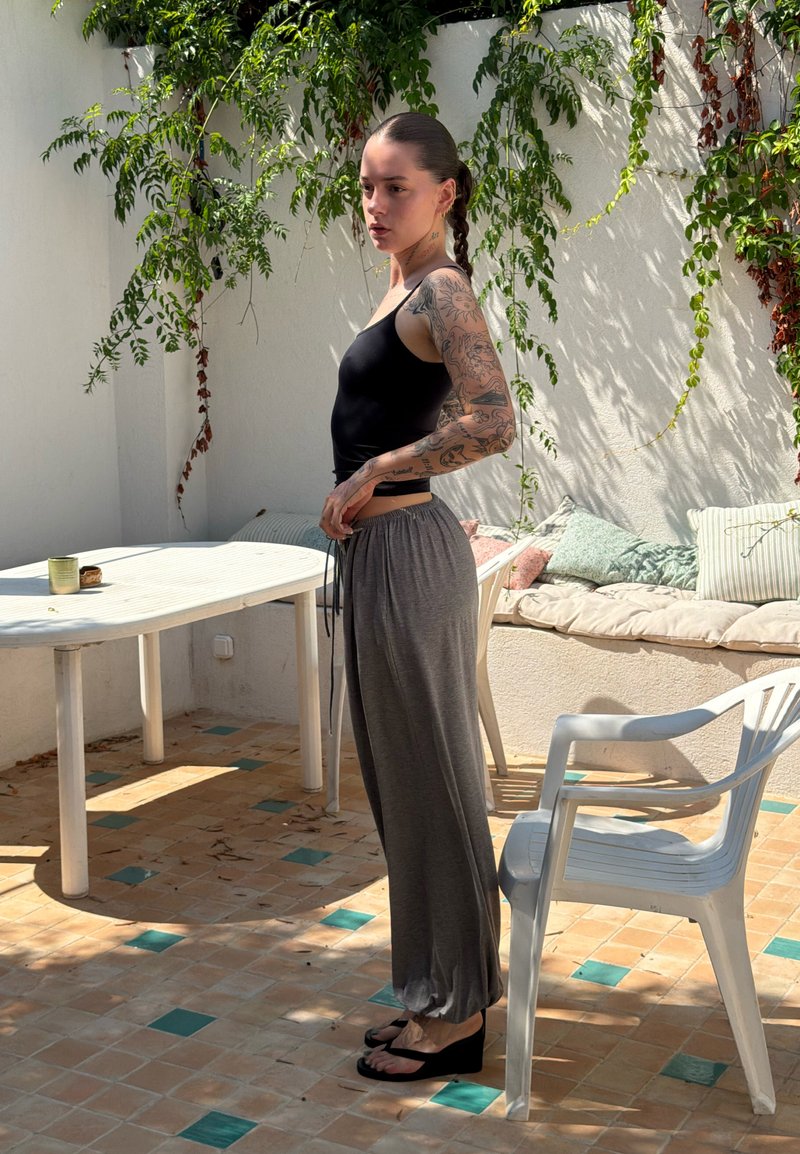 Jeune femme tatouée portant un débardeur noir et un pantalon gris se tient à côté d'une chaise et d'une table en plastique blanc sur une terrasse ensoleillée.
