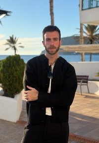 Άνδρας με κοντά σκούρα μαλλιά και γένια στέκεται με σταυρωμένα τα χέρια φορώντας μαύρη ζακέτα και λευκό πουκάμισο, με τη θάλασσα, φοίνικες και παγκάκι στο φόντο.