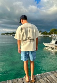 Une personne portant une casquette, un t-shirt blanc avec un motif de poisson, et un short bleu à motifs se tient sur un quai en bois surplombant une eau verte calme et des bateaux.