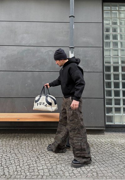 Sudadera con capucha negra, pantalones cargo de camuflaje y zapatos de plataforma negros. Lleva una bolsa de deporte de Puma en beige y negro. Fondo gris, banco de madera.
