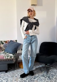 Camicia leggera a maniche lunghe, maglione nero strutturato annodato intorno al collo, jeans azzurri, pantofole nere e cappellino rosa. Ambientazione interna con divano a motivo.