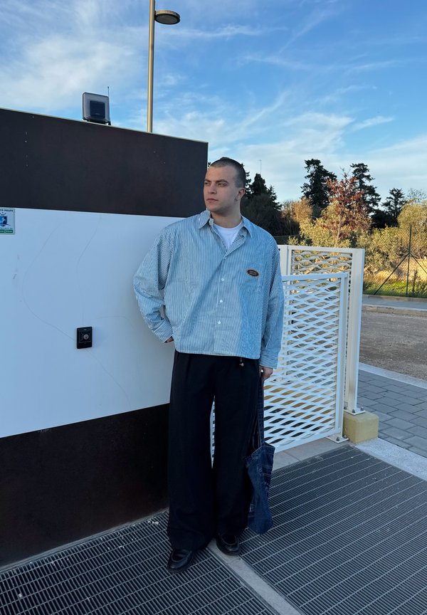 Camisa de botão riscada em azul claro e branco, ajuste solto; calças largas pretas; bolsa de mão de denim; de pé perto de uma parede branca e castanha.
