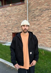 Black hooded jacket worn over an orange shirt, displaying a Carhartt logo. The individual is also wearing a cream beanie. The background features a brick wall and grass.