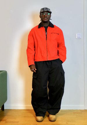 Hombre con gafas, gorra gris de Supreme, chaqueta naranja brillante, pantalones cargo negros y botas color beige, de pie en el interior frente a una pared blanca.