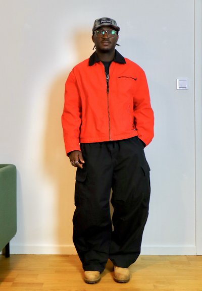 Hombre con gafas, gorra gris de Supreme, chaqueta naranja brillante, pantalones cargo negros y botas color beige, de pie en el interior frente a una pared blanca.