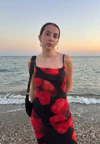 Zwarte slipdress versierd met grote rode bloemenpatronen. Het model draagt een paar grote creolen en houdt een zwarte handtas vast, terwijl ze bij de zee staat tijdens zonsondergang.