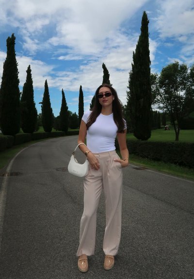 Camiseta blanca ajustada, pantalones anchos beige, mocasines color topo, acentos dorados, bolso blanco. El fondo presenta árboles altos y un cielo nublado.