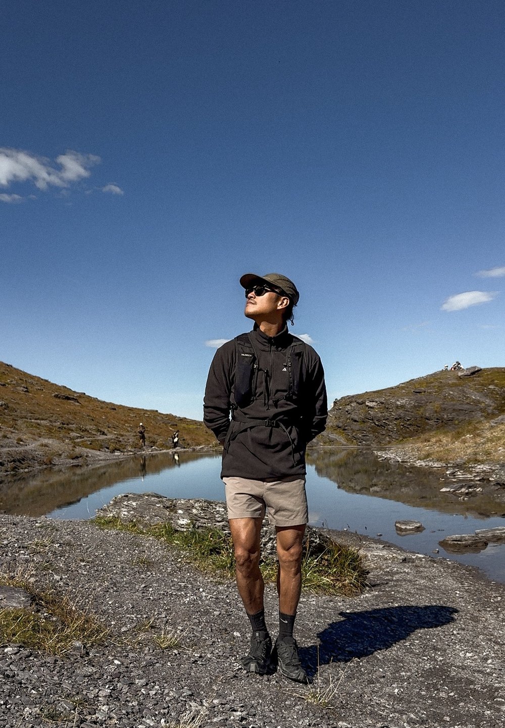 Matkasell riietatud mees seisab kivisel kaldal rahuliku järve ääres, selge sinise taeva all, taustal on künkad ja teised matkajad.