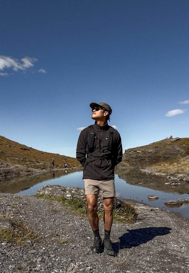Uomo in abbigliamento da trekking in piedi su una riva rocciosa vicino a un lago calmo sotto un cielo azzurro limpido, con colline e altri escursionisti sullo sfondo.