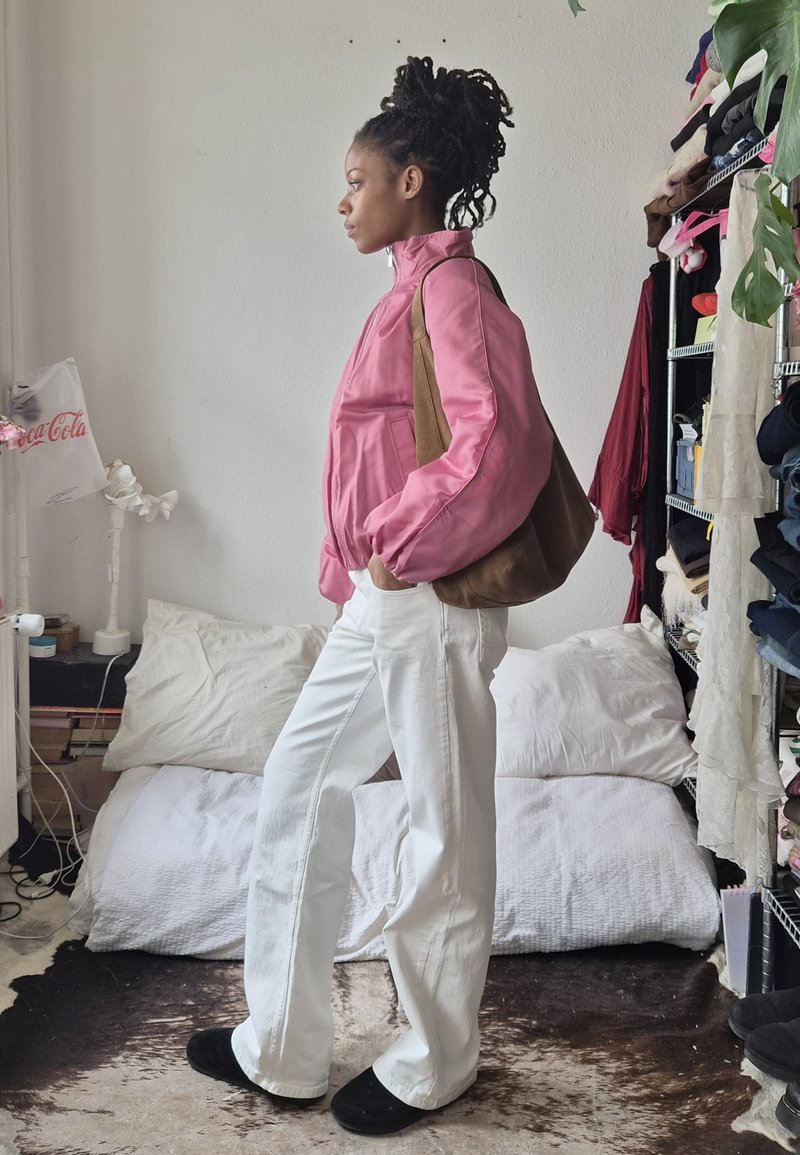 Femme avec des cheveux tressés portant une veste rose, un pantalon blanc et des chaussures noires, debout de côté en portant un sac marron dans une chambre.