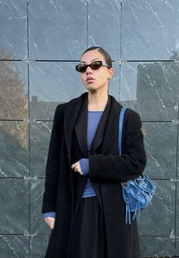 Čierny plášť, modrý top, dlhá čierna sukňa; tmavé slnečné okuliare, malá modrá kabelka; hladká látka, štruktúrovaný dizajn, minimalistické prvky.