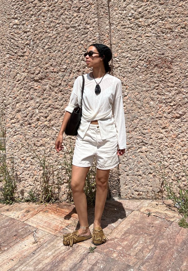 Persona con camisa blanca de manga larga y pantalones cortos, gafas de sol, zapatos marrones con borlas y llevando un bolso negro de hombro, de pie frente a una pared de piedra texturizada.