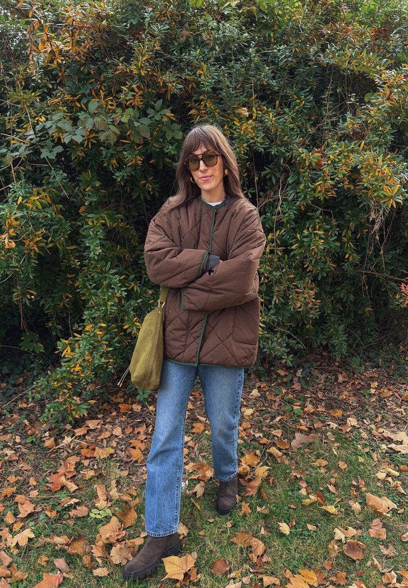 Brun quiltet oversized jakke med grønn kant, blå rette jeans, brune ankelstøvler og olivengrønn skulderveske, satt mot løvverk.