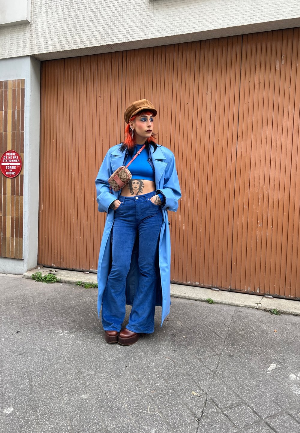 Casaco longo azul claro sobre um top cropped azul, combinado com calças de ganga azuis largas e sapatos de plataforma castanhos. Uma carteira estampada estava pendurada.
