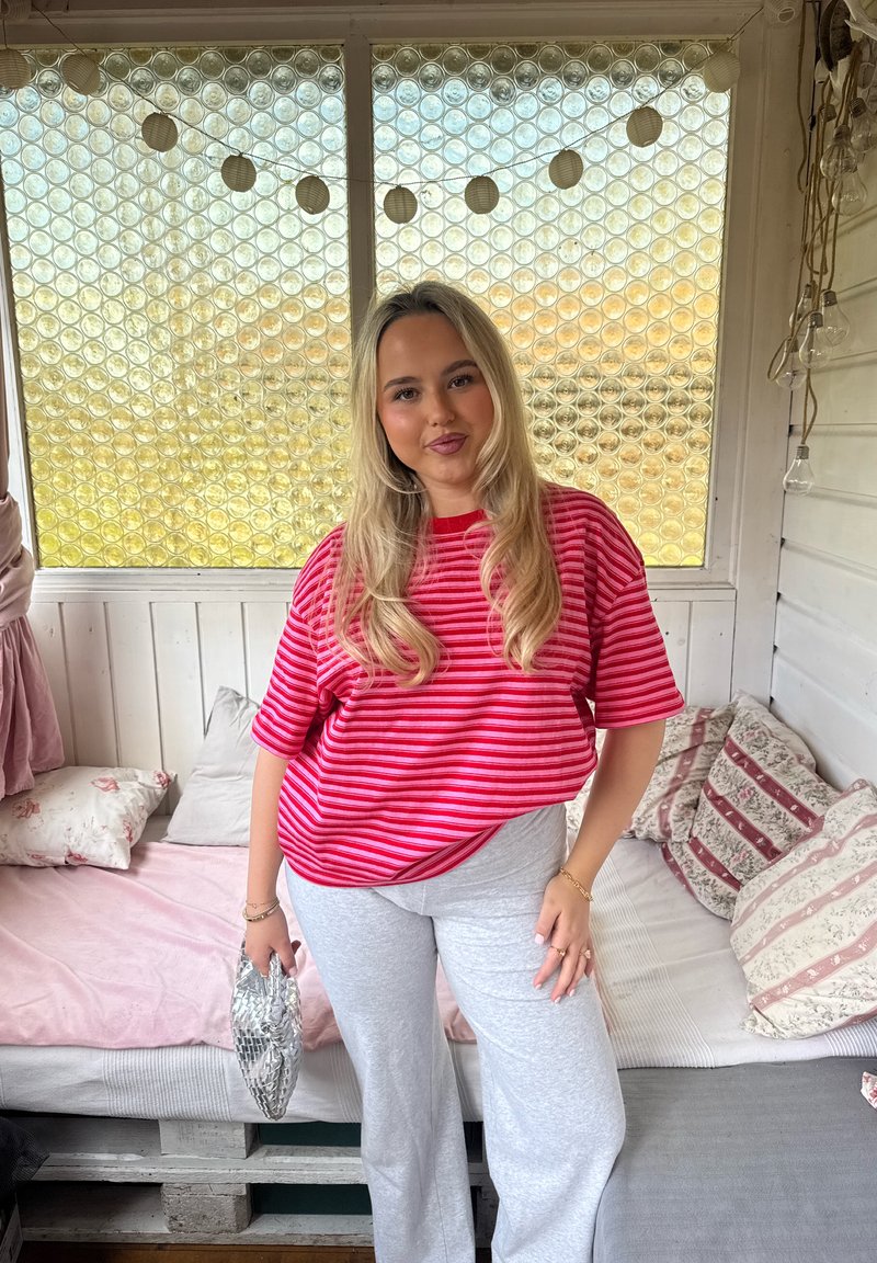 Jeune femme aux longs cheveux blonds portant une chemise rouge rayée et un pantalon gris clair, debout à l'intérieur avec des coussins décoratifs et des guirlandes lumineuses.