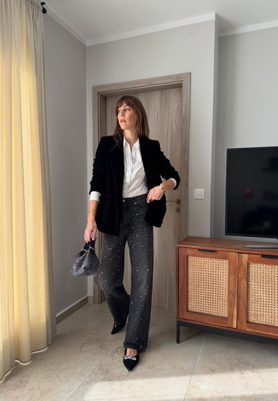 Blazer de terciopelo negro sobre una camisa blanca, combinado con vaqueros oscuros con tachuelas plateadas. Sosteniendo una bolsa de malla negra. Cortinas de color claro al fondo.