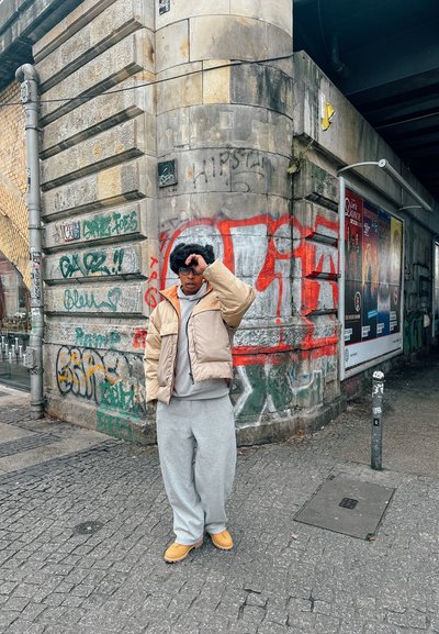 Chaqueta de plumas beige con sudadera gris debajo, pantalones de chándal grises y calzado amarillo, de pie frente a una pared de piedra cubierta de grafitis.