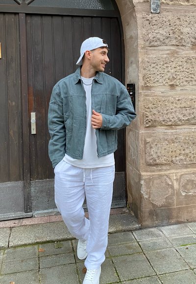 Chaqueta de mezclilla azul claro con cremallera al frente, camiseta blanca y pantalones de chándal blancos. En un fondo de pared de piedra y puerta de madera.
