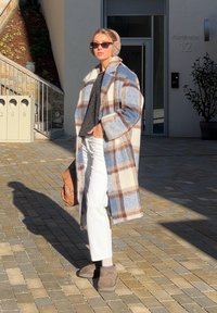 Femme debout en plein air portant un long manteau à carreaux, un pantalon blanc, des cache-oreilles beiges, des lunettes de soleil, et tenant un sac marron.