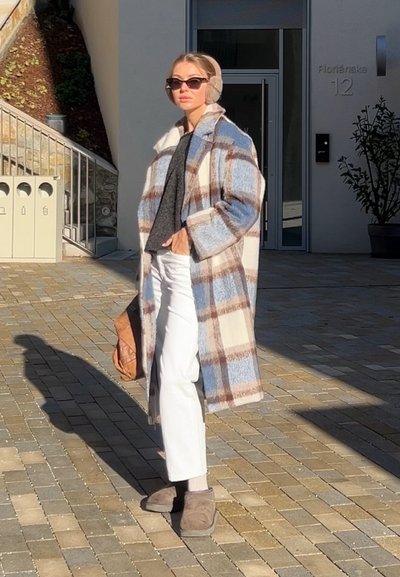 Mujer de pie al aire libre, vistiendo un abrigo largo de cuadros, pantalones blancos, orejeras beige, gafas de sol y sosteniendo un bolso marrón.