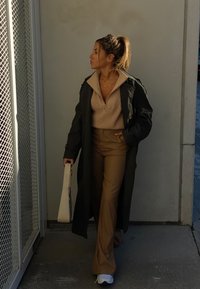 Lange zwarte jas, beige ritssweater, bruine flared broek; houdt een witte tas vast. Haar in een staart, staande tegen een lichte muur.