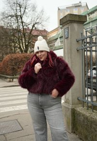 Νεαρή γυναίκα με λευκό πλεκτό καπέλο και μπουφάν από μπορντό συνθετική γούνα ποζάρει χαλαρά σε δρόμο της πόλης κοντά σε μεταλλικό φράχτη και διάβαση πεζών.