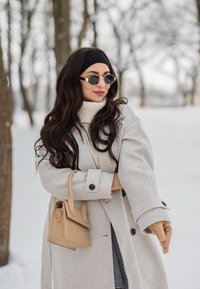 Femme en manteau d'hiver et gants, portant des lunettes de soleil et un bandeau, tient un sac à main beige en plein air dans une zone boisée enneigée.