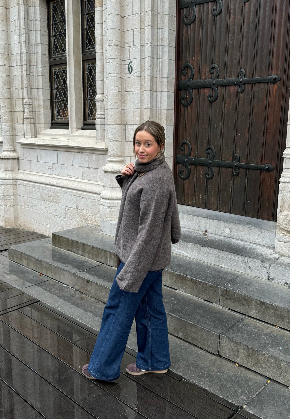 Pull en maille gris avec un col montant, associé à un jean en denim bleu ample. La scène présente un bâtiment en pierre et un pavé mouillé.