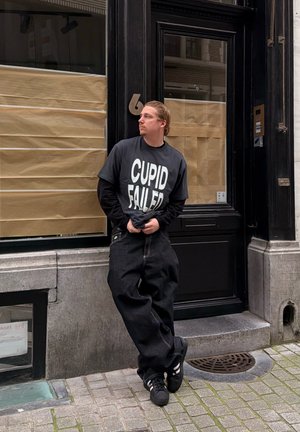 Joven con pantalones negros holgados y camiseta con la frase "CUPID FAILED" apoyado en una puerta negra en una calle empedrada.