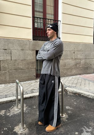 Suéter de punto gris con un dobladillo desgastado, pantalones de chándal negros con rayas blancas y botas beige. De pie al lado de un aparcamiento de bicicletas metálico.