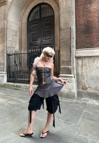 Must mustrinna, millel on lukk ette ja hall seeliku kattekiht. Mustad cargo lühikesed püksid taskutega ja nööridega; mudel kannab prille ja sandaale.