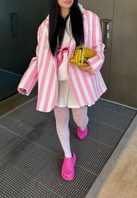Blazer de rayas rosas y blancas de gran tamaño, top blanco, crocs rosas y clutch dorado brillante. Medias blancas texturizadas completan el atuendo.