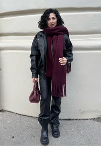 Veste en cuir noire, écharpe bordeaux avec franges, pantalon en cuir noir ajusté et bottes noires épaisses. Tenant un petit sac à main bordeaux.